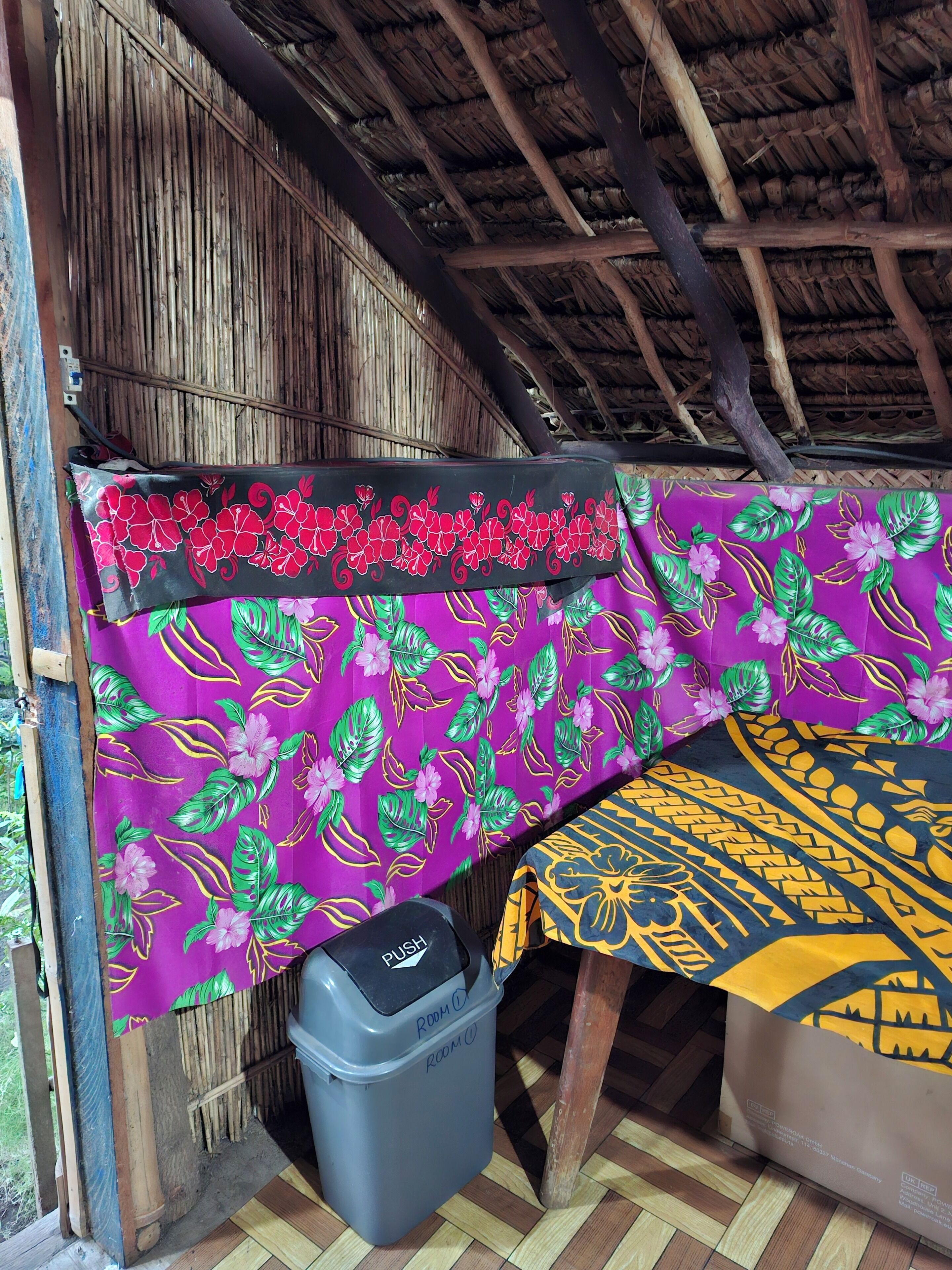 Una habitación con techo de paja, mantel de tela con estampado morado y amarillo, un cesto gris con la etiqueta 'ROOM 1' y suelo de madera.