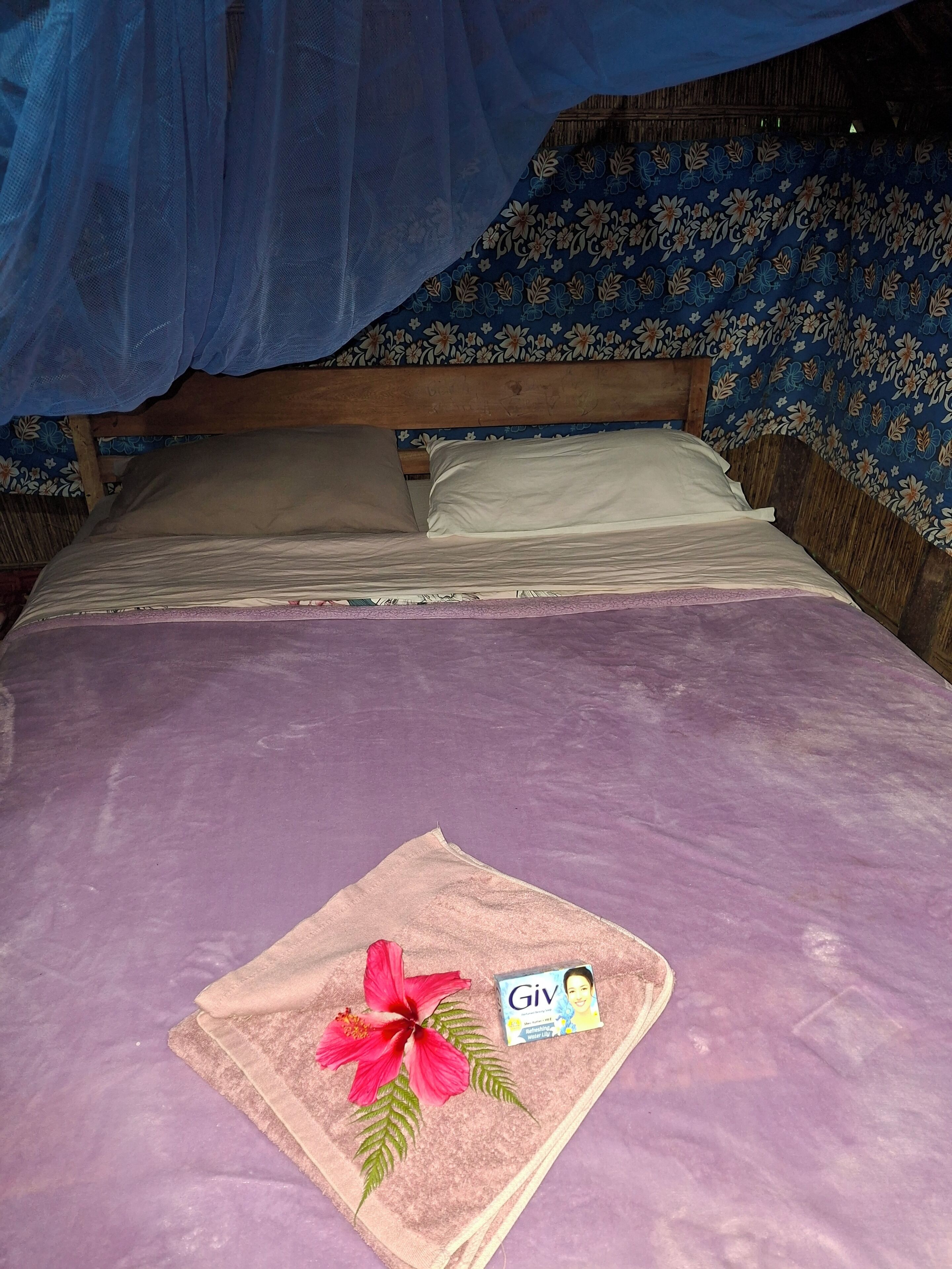 A bed with a purple bedspread, a pink towel with a hibiscus flower, and a card labeled 'Giv'.