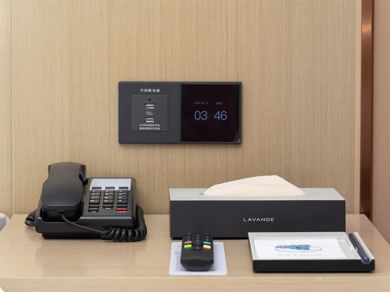 A hotel room with a digital clock displaying the time, a telephone, a remote control, and a tissue box labeled 'LAVANDE'.