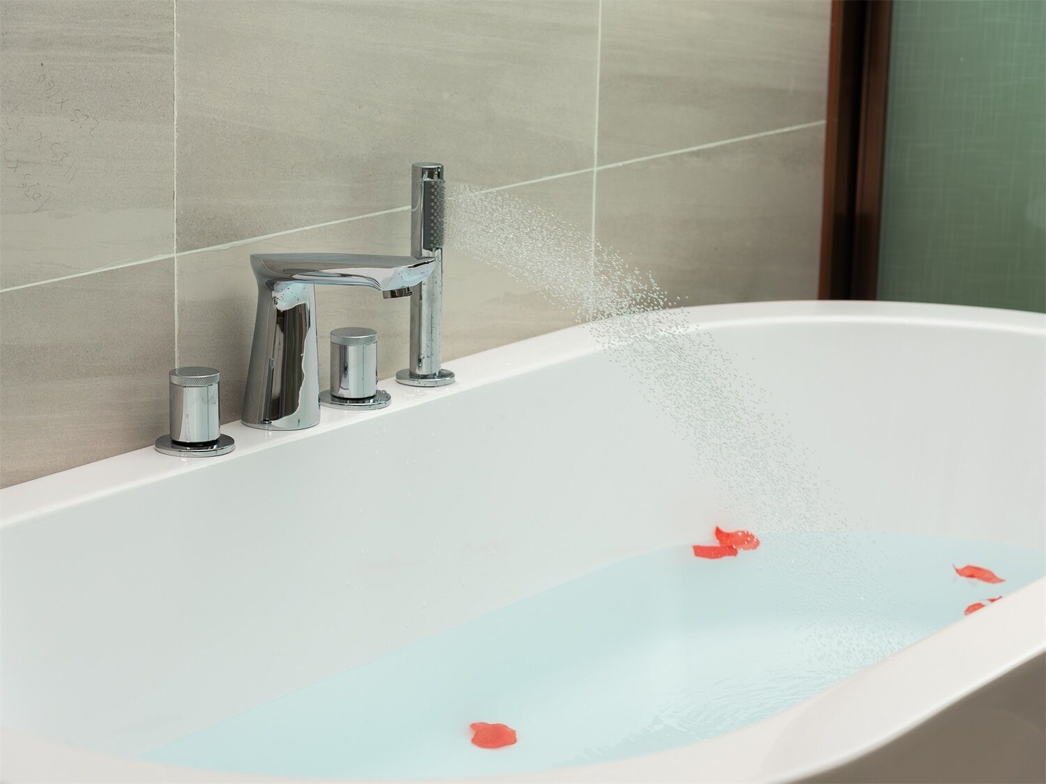 A bathtub with water flowing from a modern faucet, a small container, and a single red flower floating in the water.