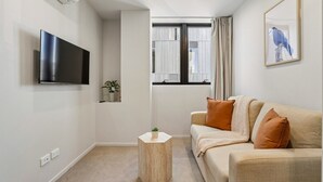 A living room with a flat-screen TV, a beige sofa with orange cushions, a small wooden side table, and a window with curtains. - Après Ski Apartment with Full Kitchen and Carpark (Queenstown)