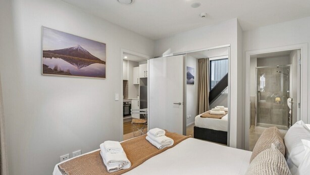 A modern hotel room with a large bed, a bathroom with a shower, and a kitchen area visible through an open door. - Après Ski Apartment with Full Kitchen and Carpark (Queenstown)