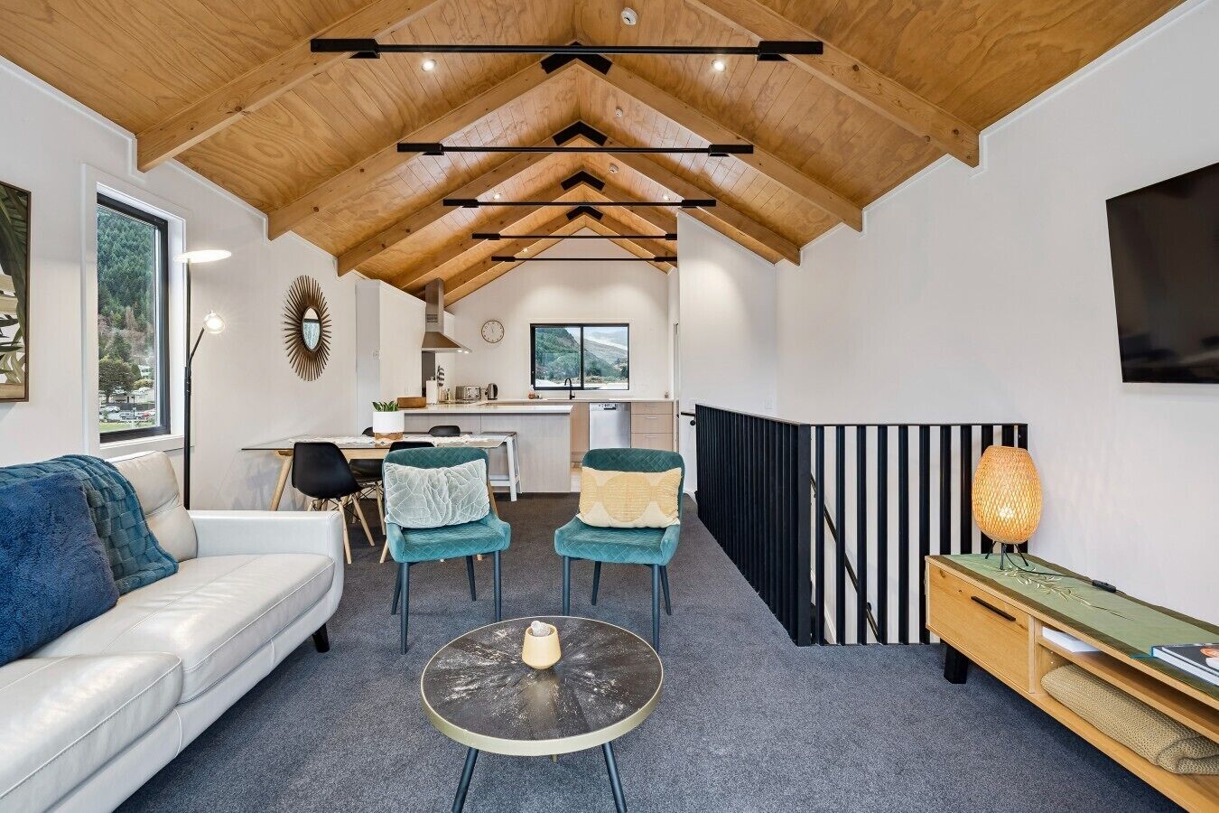 A modern interior with a high ceiling, a sofa, two chairs, a small round table, a kitchen area, and a TV mounted on the wall.