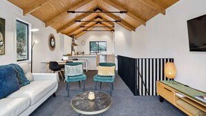 Un interior moderno con techo alto, un sofá, dos sillas, una mesita redonda, una zona de cocina y un televisor montado en la pared.