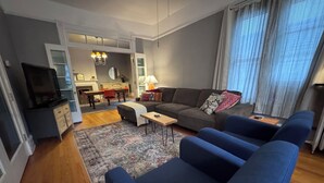 Ein modernes Wohnzimmer mit einer Couch, einem Couchtisch und einem Fernseher. Es gibt einen Essbereich mit Tisch und Stühlen. Der Raum verfügt über ein großes Fenster mit Vorhängen.