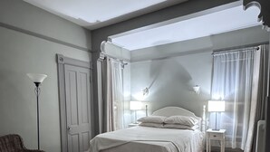 Ein Schlafzimmer mit Bett, Sessel, Nachttisch, Lampe und einem Fenster mit Vorhängen.
