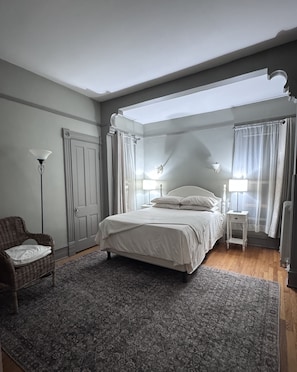 A bedroom with a bed, a chair, a nightstand, a lamp, and a window with curtains.