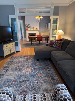 A living room with a sofa, ottoman, TV, fireplace, dining table, chairs, and a patterned rug.