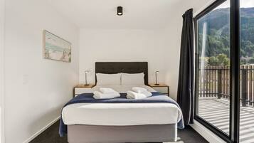 A hotel room with a bed, two bedside tables, a painting on the wall, and a large window with a view of the outdoors.