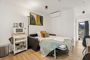 A compact hotel room with a bed, a kitchenette, a small table, and a view of a balcony. - Alpine Vista Apartment. Easy Walk to CBD! (Queenstown)