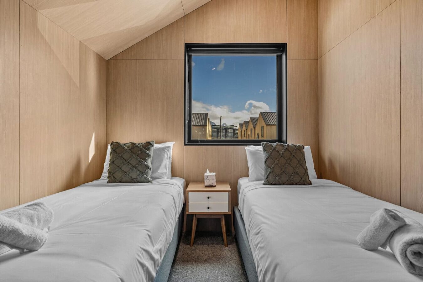 Una habitación pequeña y moderna con dos camas, una mesita de noche, un ventanal con vistas a edificios y paredes de madera.