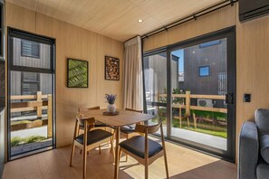 Une pièce moderne avec une table à manger en bois, des chaises, une plante en pot et de grandes fenêtres donnant sur une terrasse en bois et un bâtiment.