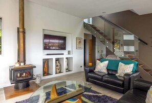 A modern living room with a glass coffee table, a black leather sofa, a wooden fireplace, and a staircase with glass railings. - Paradise at Palm Beach (Waiheke Island)