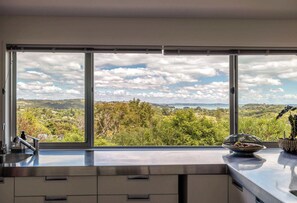 A kitchen with a large window offering a view of a landscape with trees and hills. - Paradise at Palm Beach (Waiheke Island)