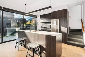 Cozinha moderna com ilha central, dois banquetas altas, geladeira de aço inoxidável e vista através de grandes portas de vidro.