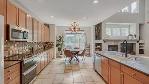 A modern kitchen with wooden cabinets, a dining area with a chandelier, and a large window. - Comfy 4-Bedroom House in Castle Rock with Priv. Fenced-in Backyard/Dog Friendly. (Castle Rock)
