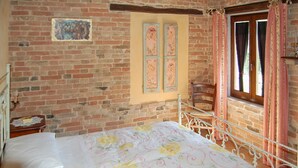 A room with a brick wall, a bed with floral bedding, a wooden chair, and a window with curtains. - Lovely home in Sarnano with swimming pool (Sarnano)