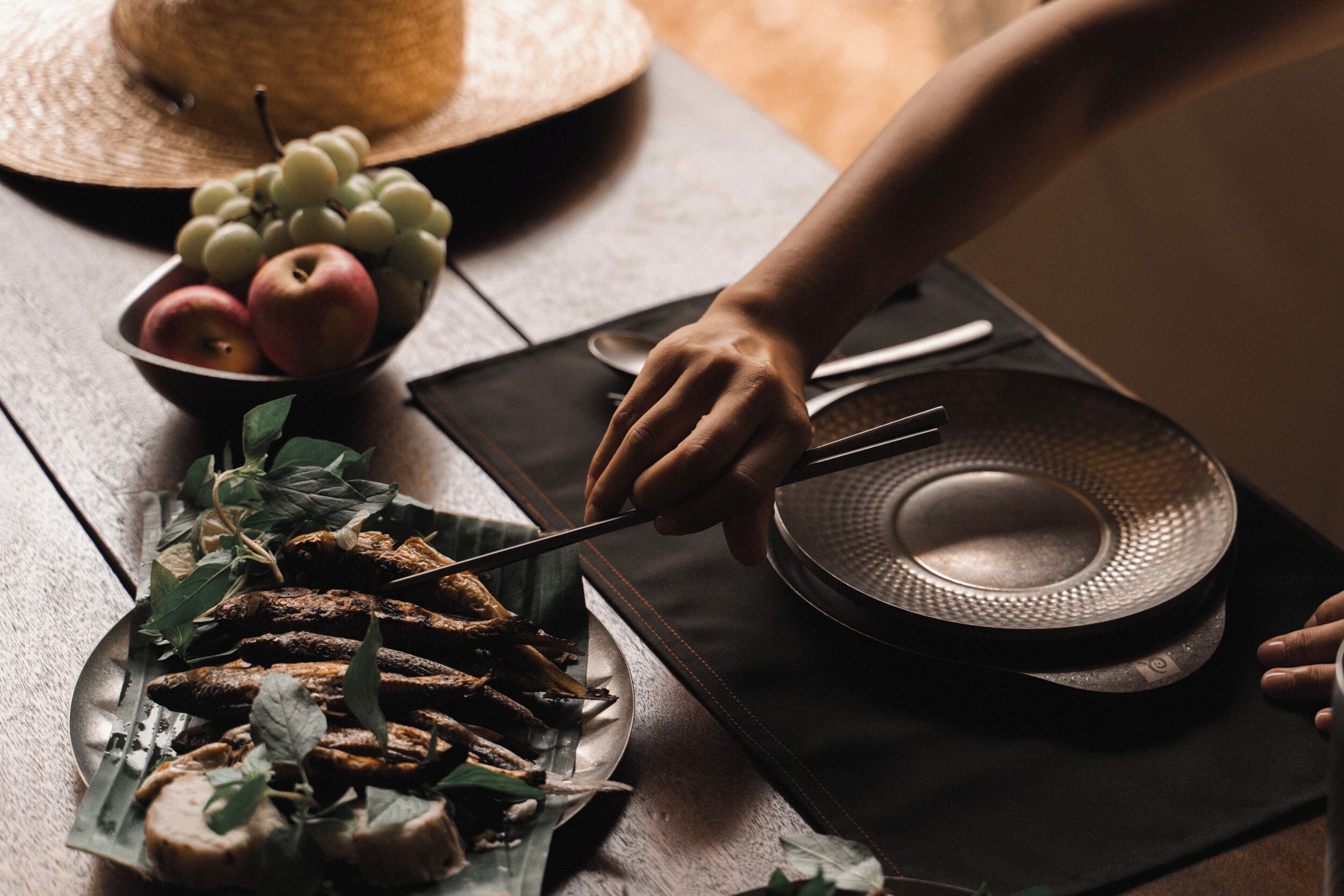 A dining table with a plate of sliced bread, a bowl of fruit, and a person cutting bread.