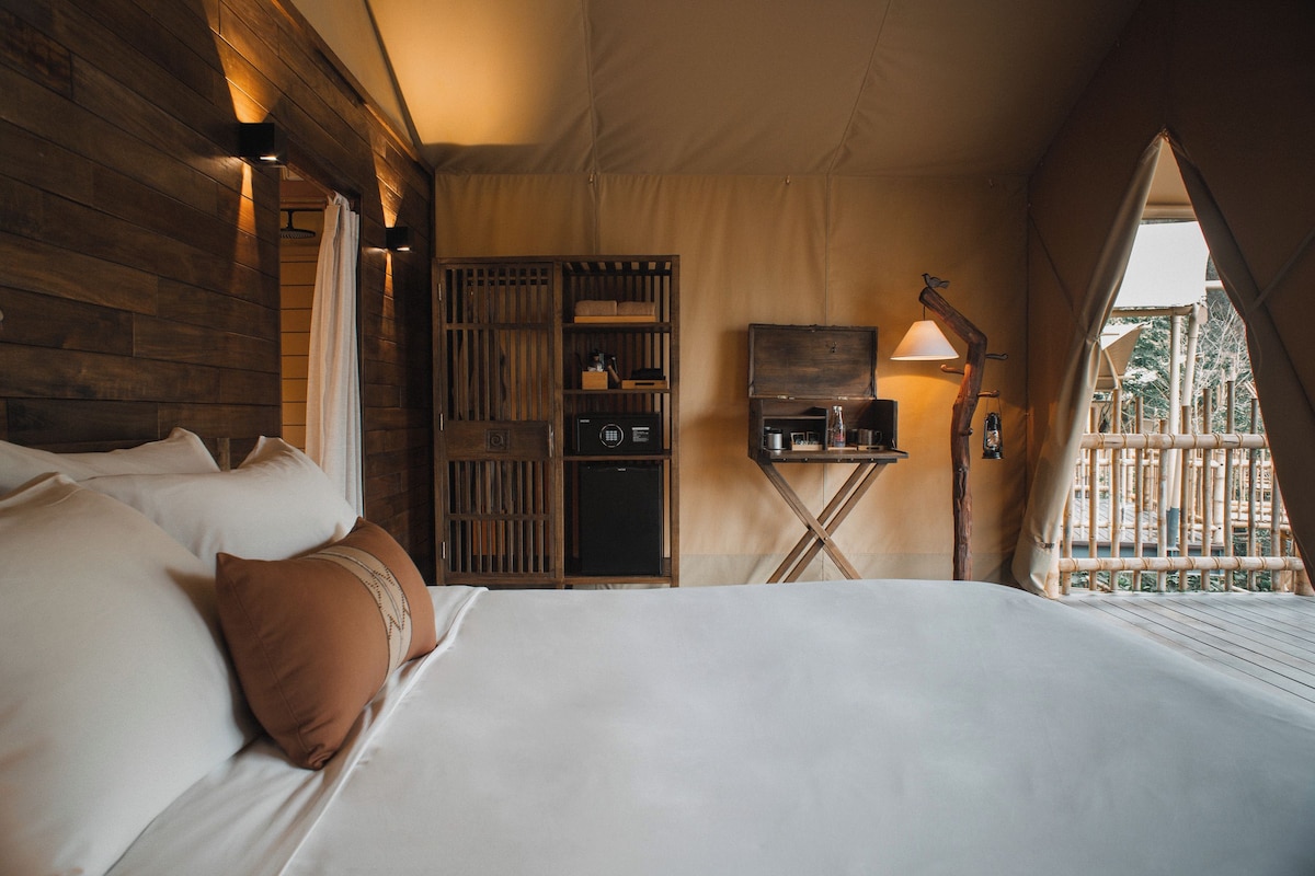 Un dormitorio con cama, almohadas, un pequeño armario de madera, una lámpara y una ventana con vistas.