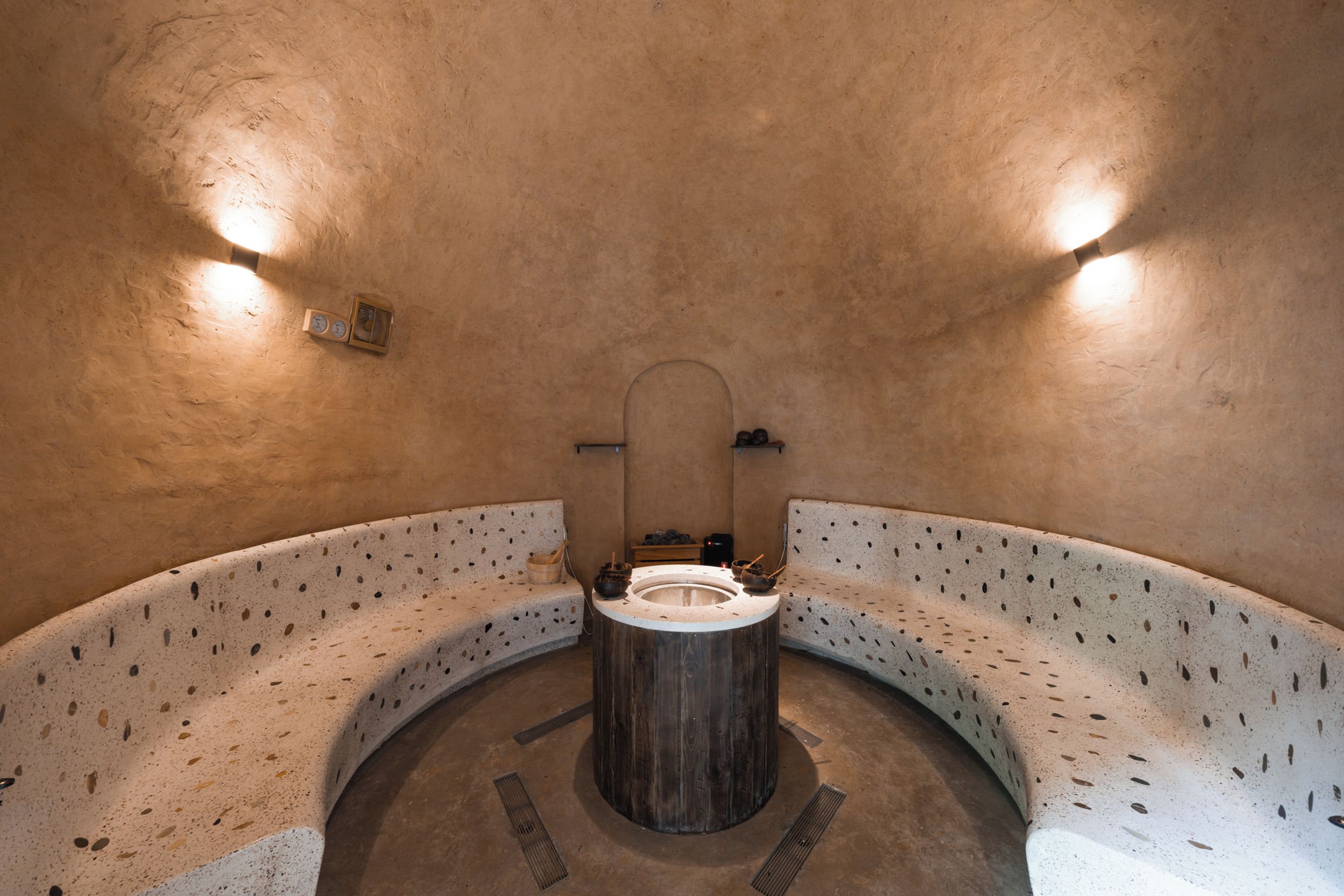 Sauna circular de paredes de piedra con un banco de madera y una estructura central de madera, posiblemente un asiento o calentador.