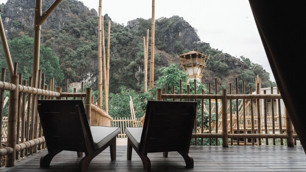 A wooden deck with two lounge chairs, bamboo railings, and a view of a mountainous landscape.