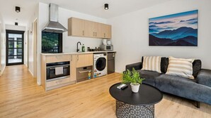 A modern kitchen with a stove, oven, microwave, and washing machine. There is a sofa with cushions and a potted plant on a coffee table.