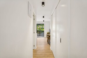 Un couloir étroit aux murs blancs et au plancher en bois mène à un balcon aménagé avec du mobilier d’extérieur.