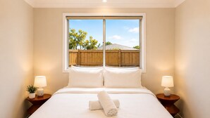 Una cama bien hecha con sábanas blancas, dos lámparas de noche y una ventana que da a una valla de madera y árboles.