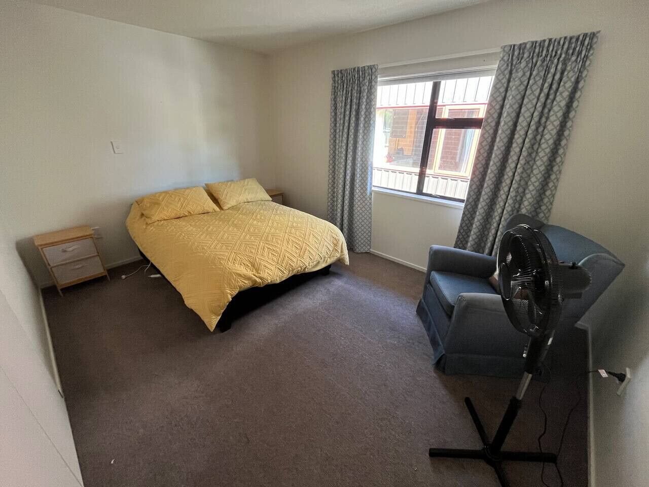 A small, sparsely furnished room with a bed, a desk, a chair, and a standing fan.