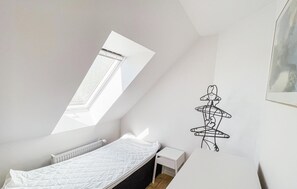 Une petite chambre à coucher aux murs blancs, avec un toit en pente, une lucarne, un lit simple, une petite table de chevet et un porte-manteau fixé au mur.