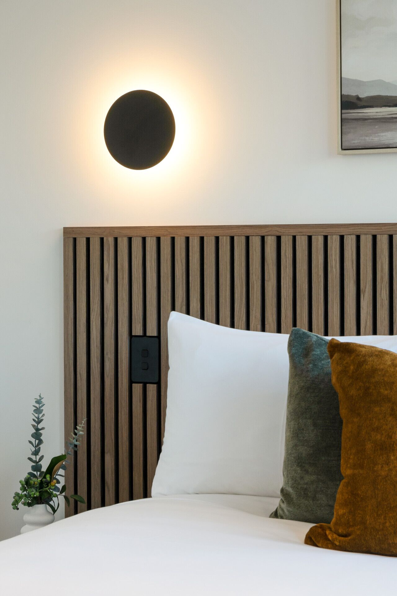 A bed with white bedding, a wooden slatted headboard, a round wall light, and a small plant.