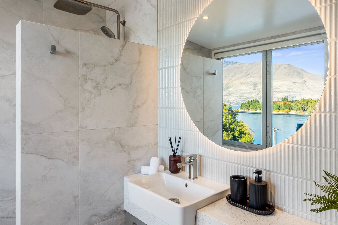 A bathroom with a shower, a sink, a mirror, and a view of a lake and mountains.