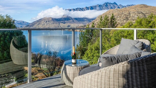 Ein Balkon mit Korbmöbeln, einer Champagnerflasche und einem Glas auf einem Tisch, mit Blick auf einen See und Berge.