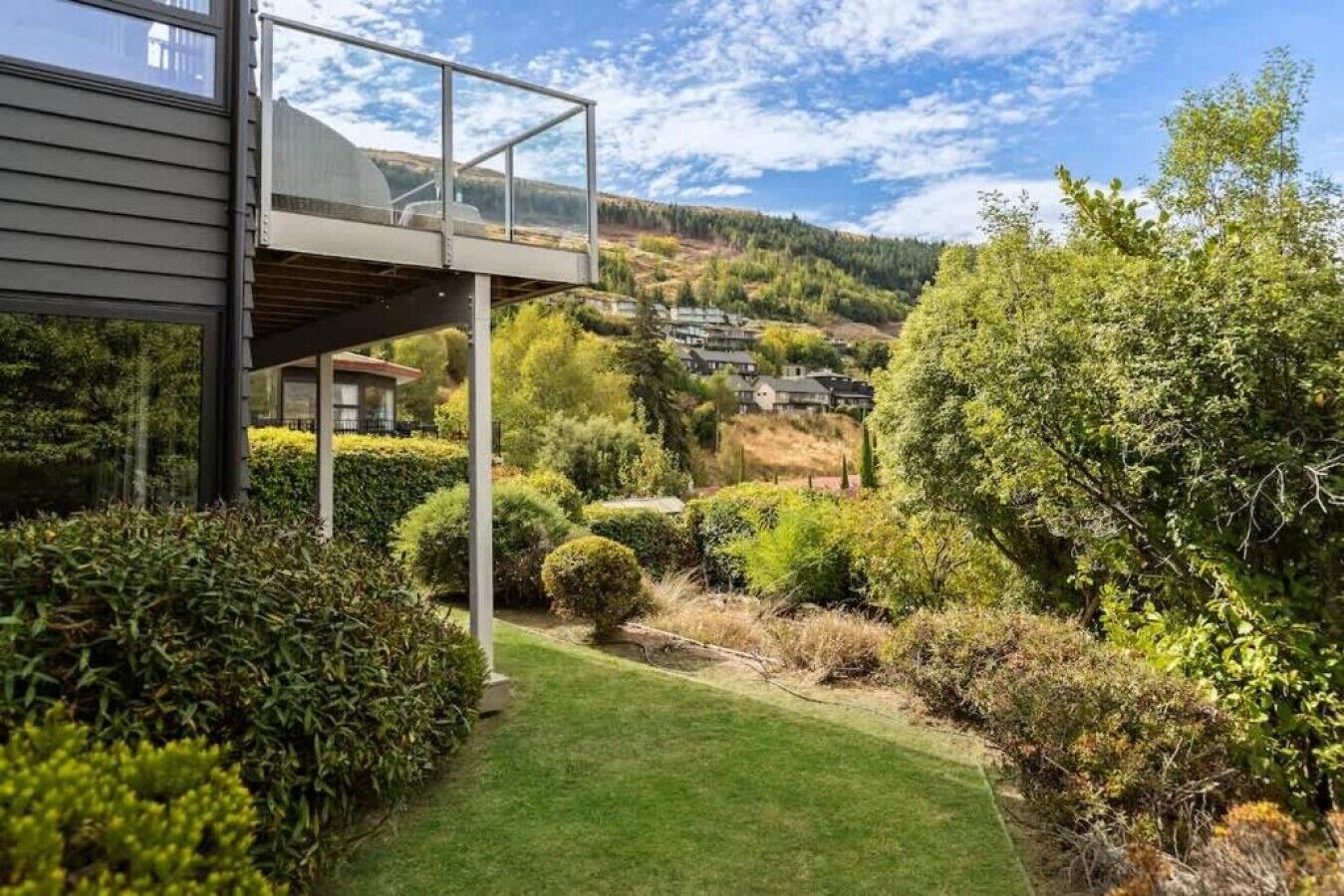 A modern house with a glass balcony overlooking a well-maintained garden and a hilly landscape in the background.