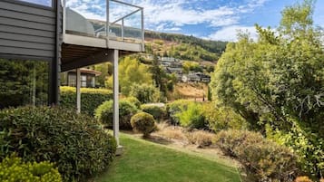 A modern house with a glass balcony overlooking a well-maintained garden and a hilly landscape in the background.