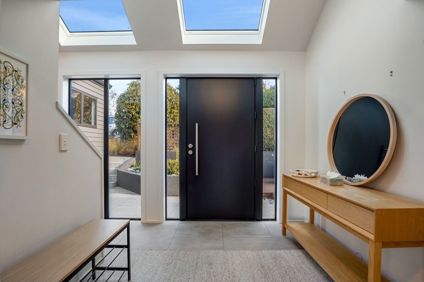 A modern interior with skylights, a black door, a wooden bench, and a round mirror on a wooden console.