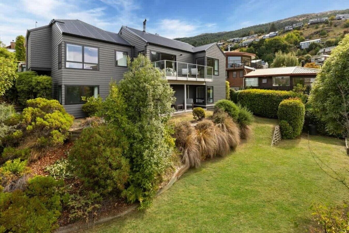 Une maison moderne à deux étages, dotée d’un balcon, entourée de jardins bien entretenus et bordée par un paysage vallonné.