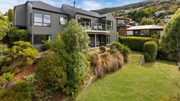 Une maison moderne à deux étages, dotée d’un balcon, entourée de jardins bien entretenus et bordée par un paysage vallonné.