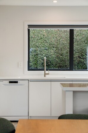 A modern kitchen with a built-in dishwasher, a large window with a view of a hedge, and a wooden table. - The Gold Rise | Two Living Areas + Lake Views (Queenstown)