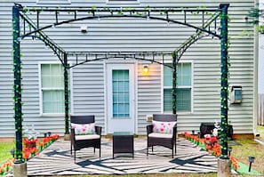 A covered patio with two chairs and a table, a house with a porch, and a garden with red flowers. - D's Creekside NorthCarolinaHome Apartment A (Burlington)