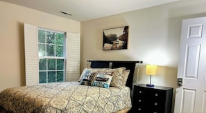 A bedroom with a bed, a nightstand, a lamp, a window with shutters, and a painting on the wall.