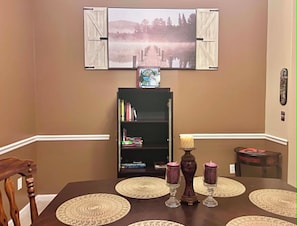 A dining area with a table set for two, a bookshelf, and a mounted television displaying a scenic image.