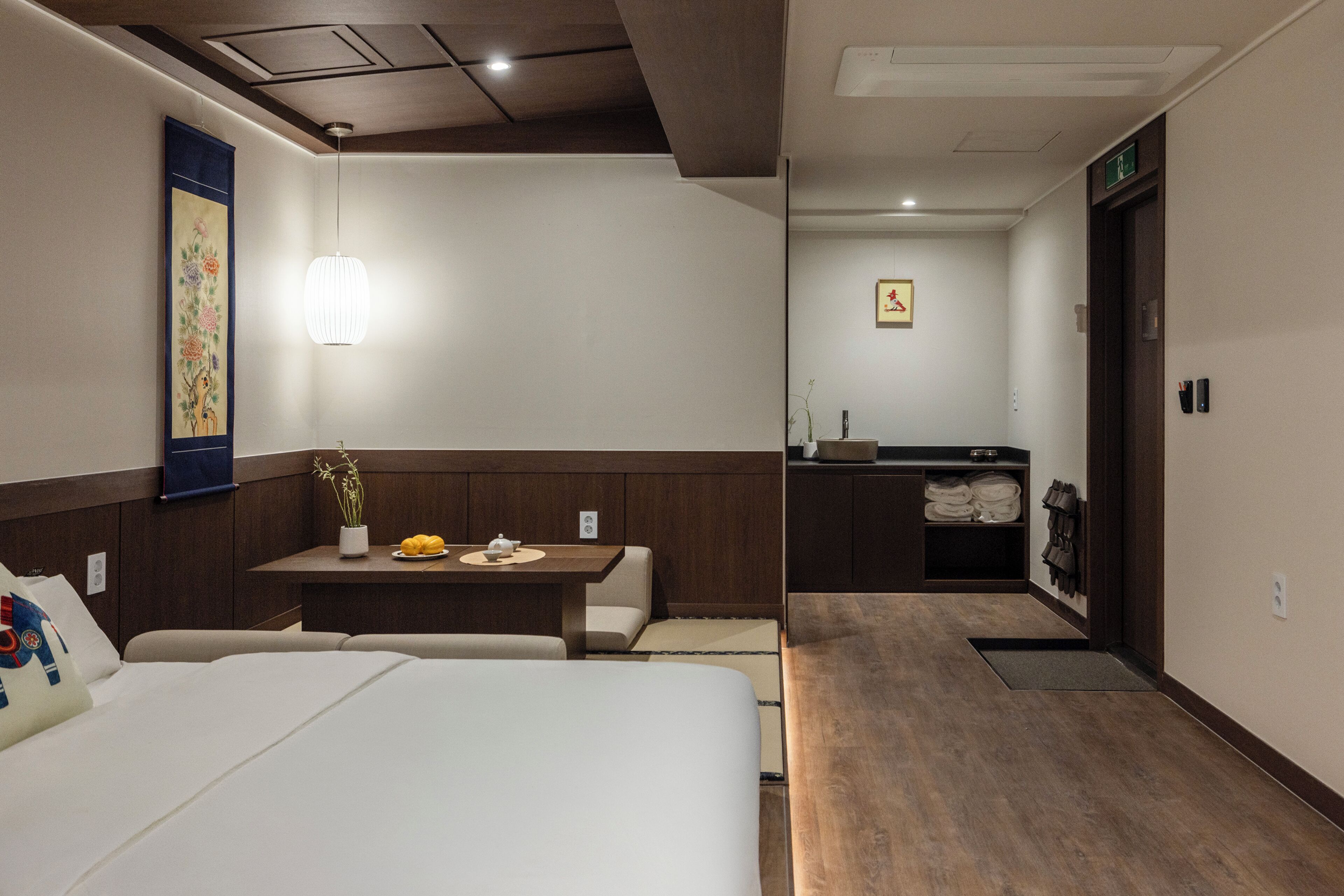 A hotel room with a bed, a small table with a vase and fruit, a bench, a wall-mounted artwork, a wooden cabinet, and a door with an exit sign.