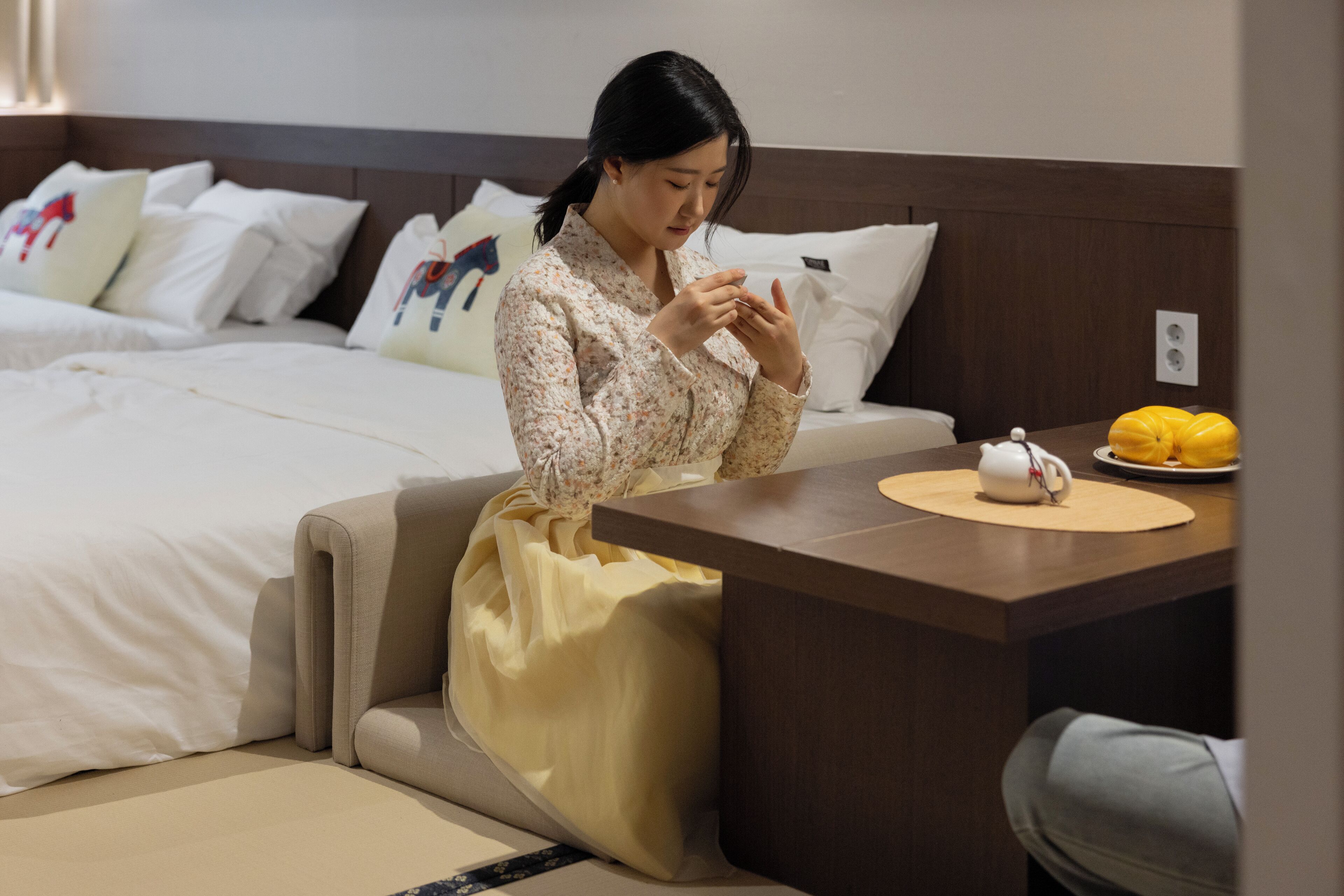 A hotel room with a bed, a bedside table with a cup and a plate of fruit, and a sofa.