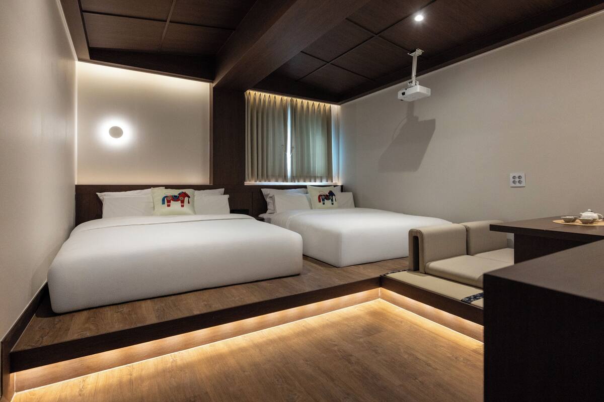 Una habitación de hotel moderna con dos camas, un sofá, suelo de madera y un proyector en el techo.
