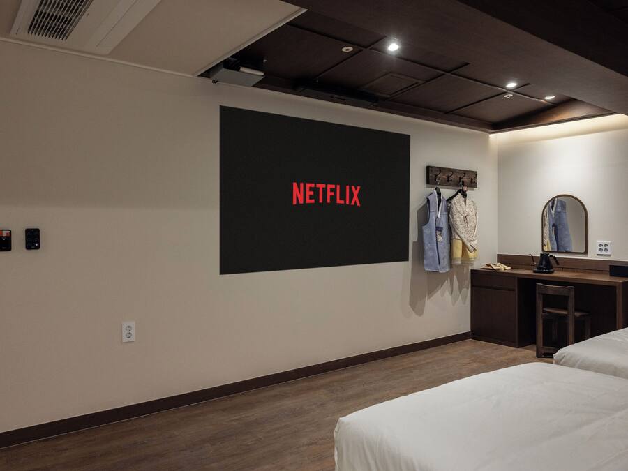 A hotel room with a large screen displaying 'NETFLIX,' a desk with a mirror, and two beds.