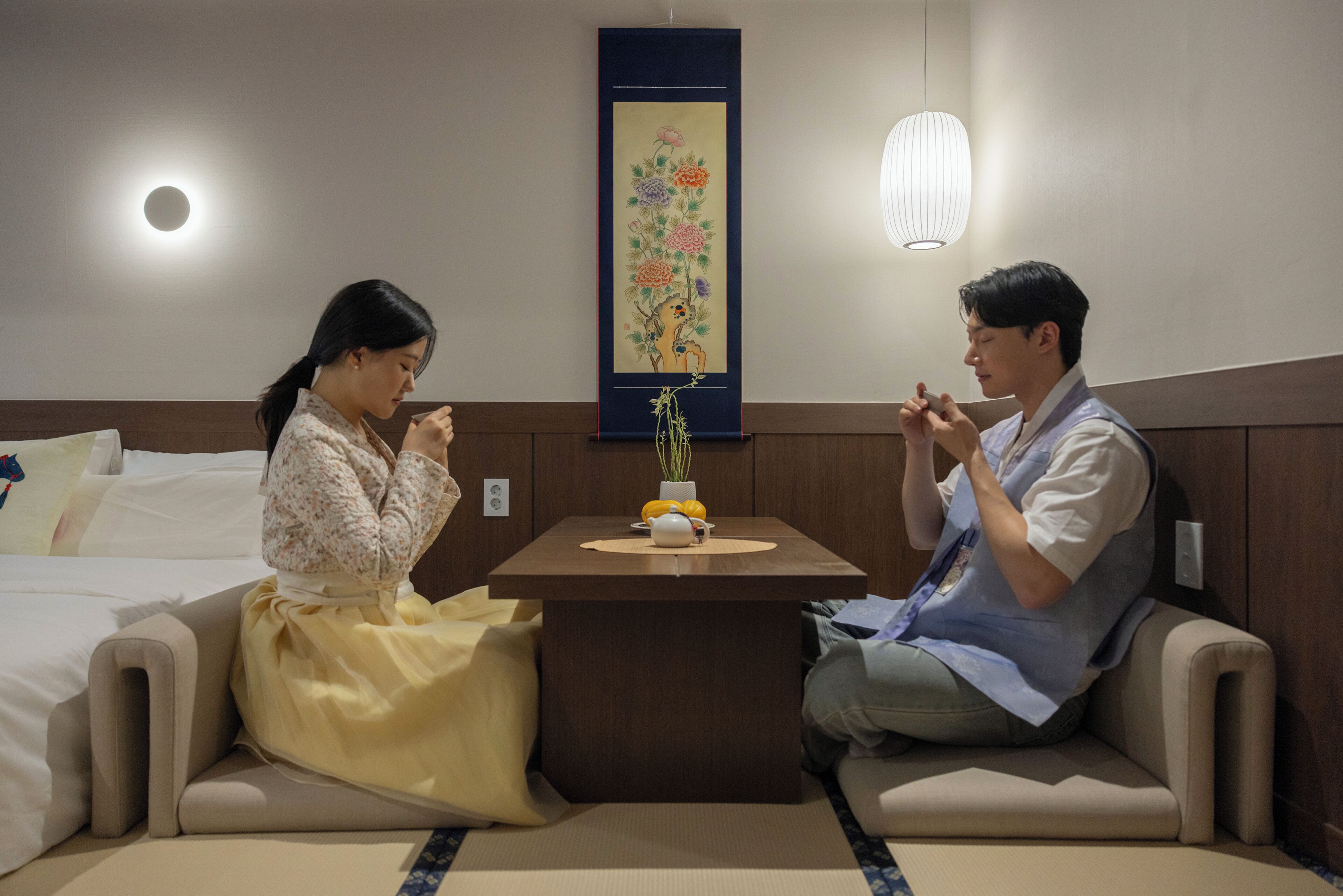 A room with a bed, a small table with a vase and a teapot, and a decorative painting on the wall.