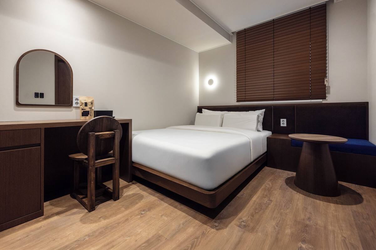 Habitación de hotel con cama, escritorio, silla, una mesa redonda y ventana con persianas.