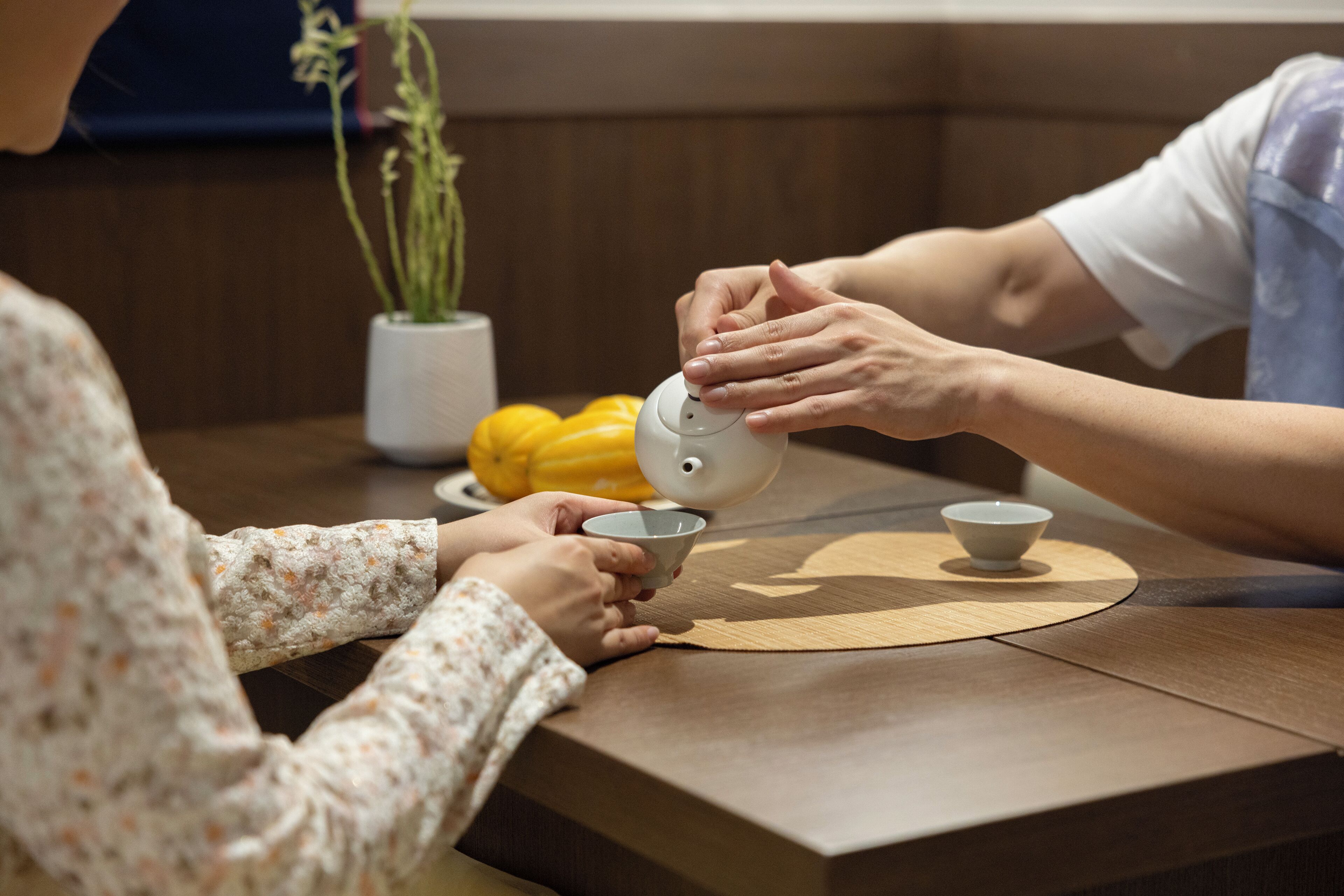 A person pouring tea into a cup at a wooden table with a plant in the background.