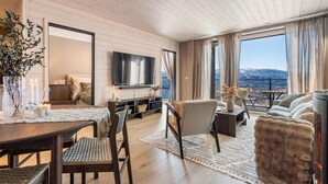 Ein modernes Wohnzimmer mit Flachbildfernseher, Holzboden, einer Essecke mit Tisch und Stühlen sowie einem großen Fenster mit Blick auf die Berge.